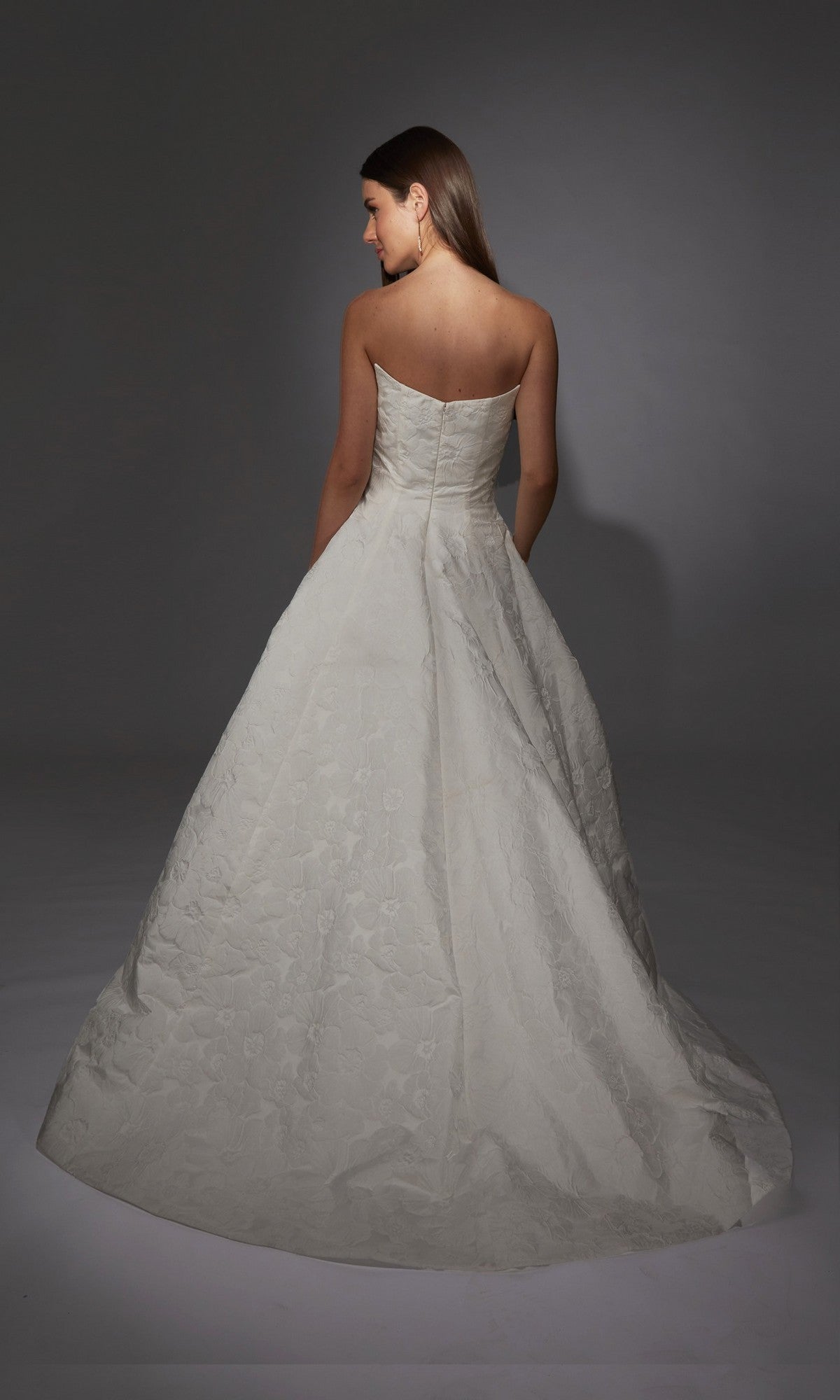 A woman wearing the Alyce 7155 long white strapless gown with elegant floral jacquard fabric stands facing away from the camera against a plain gray background.